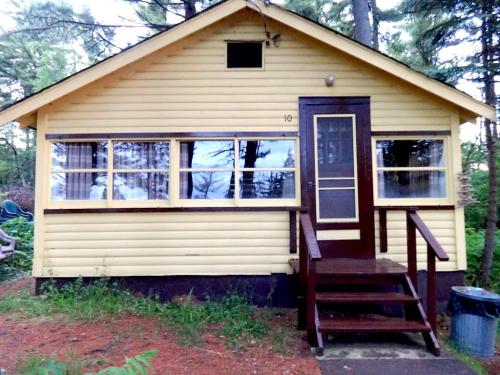 A yellow cabin with a wooden porch, surrounded by trees and greenery, featuring large windows and a simple design.