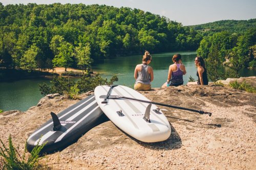 Three women sit on a rock by a lake, with two paddleboards resting nearby and lush greenery in the background.