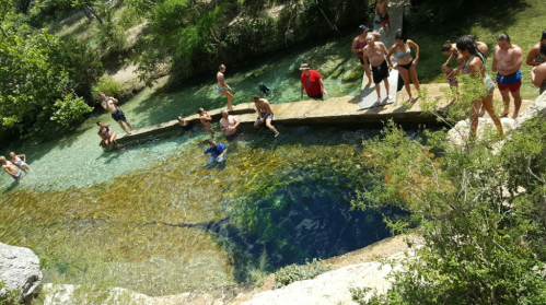 A group of people enjoying a natural swimming area with clear water surrounded by greenery on a sunny day.