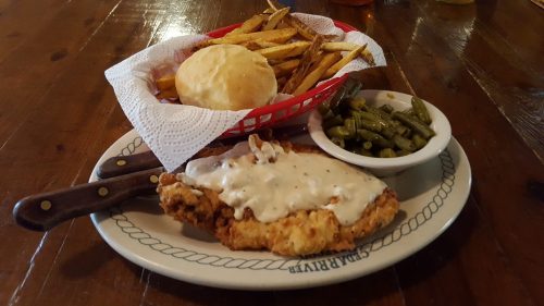 A plate of fried chicken smothered in gravy, served with fries, green beans, and a biscuit on a wooden table.
