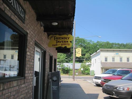 Sign for "Joe's Friendly Tavern" hangs above a brick building, with a clear blue sky and trees in the background.