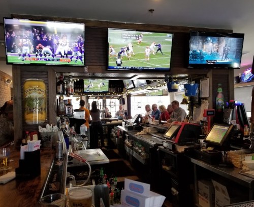 A lively bar scene with multiple TVs showing sports, a well-stocked bar, and patrons enjoying drinks and food.