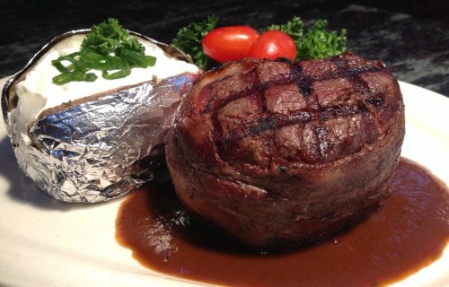 A grilled steak with a baked potato topped with sour cream and green onions, garnished with cherry tomatoes and parsley.