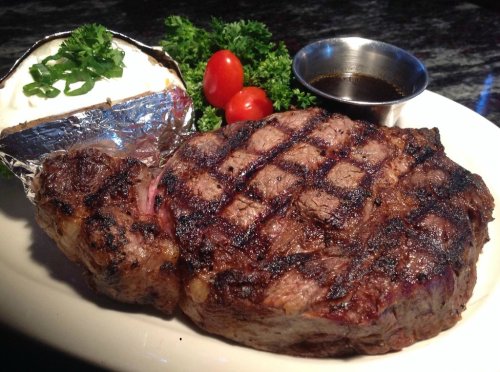 A grilled steak with a crosshatch pattern, served with a baked potato, cherry tomatoes, and a small bowl of sauce.