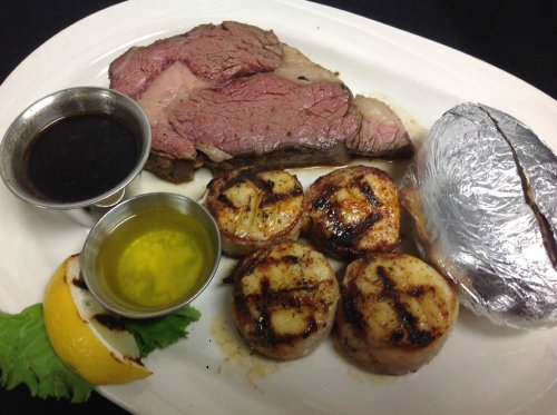 A plate featuring a slice of roast beef, grilled scallops, a baked potato, and sauces, garnished with lemon and lettuce.