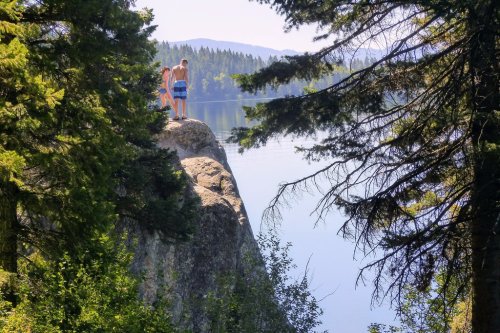 Two people stand on a rocky ledge by a calm lake, surrounded by trees and mountains in the background.