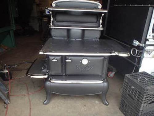 A vintage black cast iron stove with a shiny metal trim and multiple compartments, set against a workshop background.
