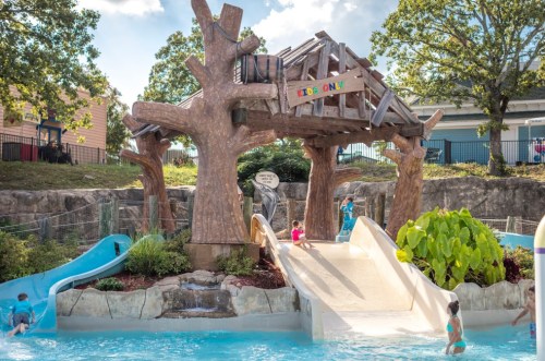 A children's water play area featuring slides, a wooden structure, and lush greenery under a sunny sky.