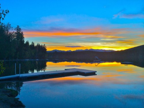 A serene lake at sunset, reflecting vibrant colors in the sky and surrounding trees, with a dock in the foreground.