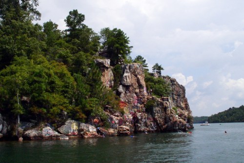 A rocky cliff covered in greenery rises from a calm lake, with people swimming nearby under a cloudy sky.