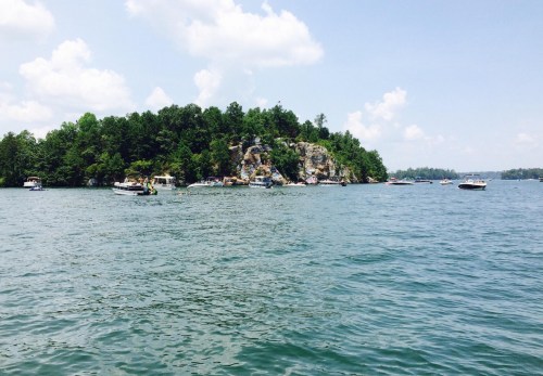 A scenic view of a rocky island surrounded by water, with boats and lush greenery under a bright blue sky.