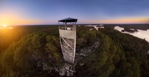 A panoramic view of a fire tower overlooking a forested landscape at sunset, with lakes in the distance.
