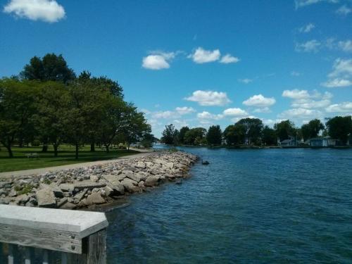 A serene waterfront scene with a rocky shoreline, lush trees, and a clear blue sky dotted with fluffy clouds.