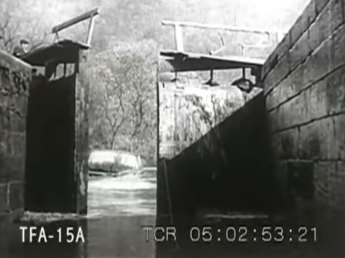 A black and white image of a canal lock with gates partially open, surrounded by trees and water.