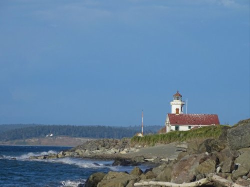 A lighthouse stands on a rocky shore, with waves crashing and a distant forested island under a blue sky.