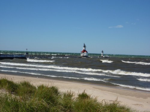 A scenic view of a lighthouse on a pier, with waves crashing and sandy beach in the foreground under a clear blue sky.