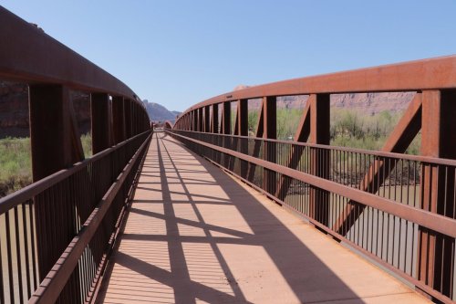 A rust-colored metal bridge spans a river, with shadows cast on the walkway and greenery visible on either side.