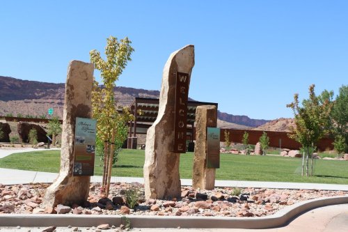 Welcome sign made of stone pillars in a landscaped area with greenery and red rock formations in the background.