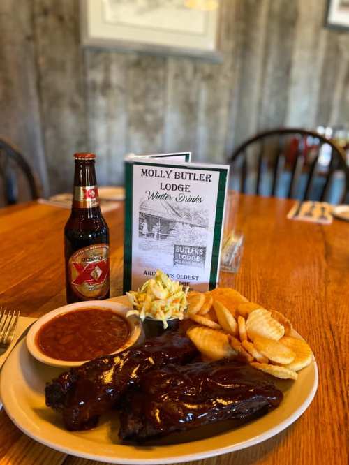 A plate of BBQ ribs with baked beans, coleslaw, and fries, alongside a bottle of soda and a menu on a wooden table.