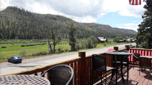 A scenic view from a wooden deck overlooking a green valley and mountains, with a car parked on the road below.