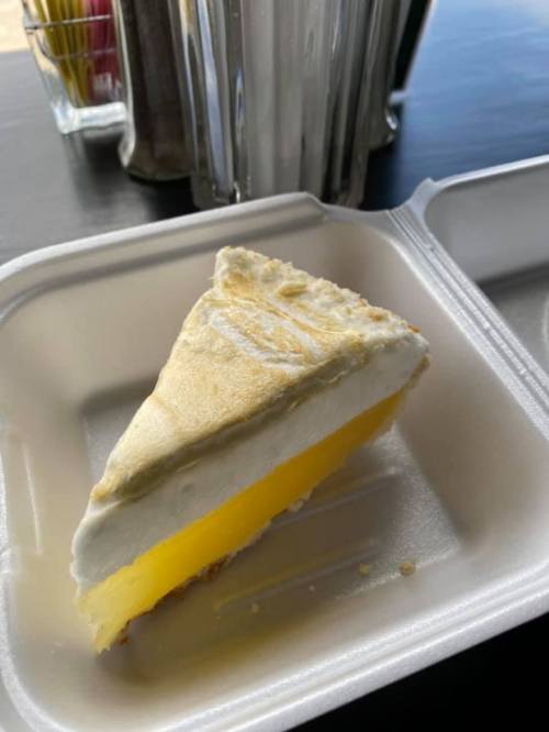 A slice of lemon meringue pie in a takeout container, featuring a yellow filling and fluffy white topping.