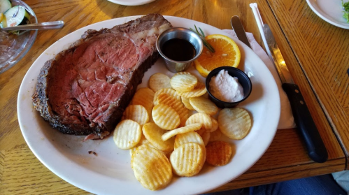 A plate with a large slice of prime rib, dipping sauce, orange slice, and a side of crispy potato chips.