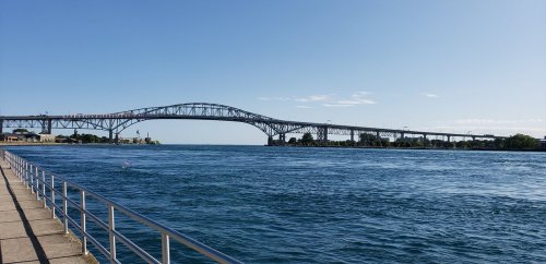 A wide river with a large bridge spanning across it under a clear blue sky. A walkway runs along the riverbank.