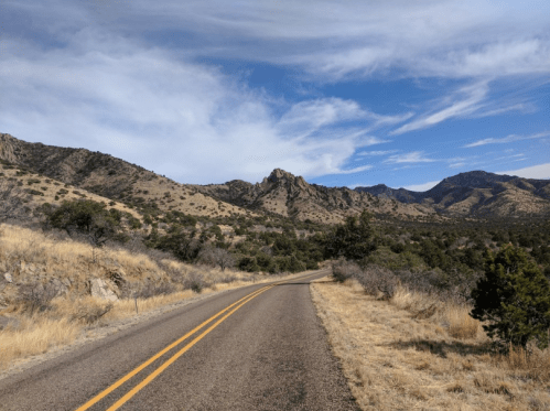 A winding road through dry grasslands and mountains under a blue sky with scattered clouds.