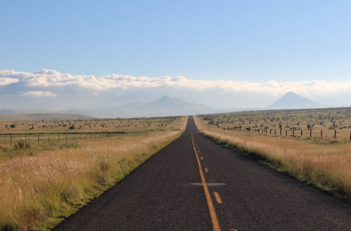 A long, empty road stretches through grassy fields, leading towards distant mountains under a clear blue sky.