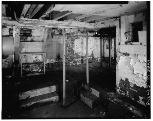 Black and white image of a dimly lit basement with exposed beams, wooden stairs, and weathered walls.