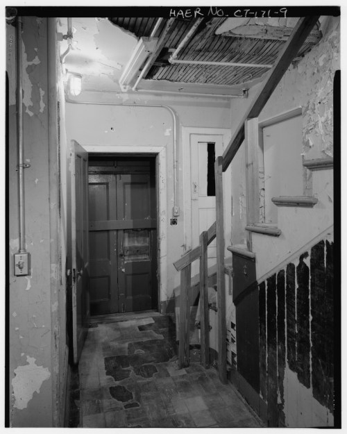 Black and white image of a worn hallway with stairs, peeling paint, and a closed door at the end.