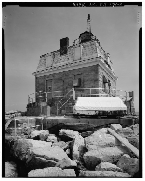 A historic lighthouse made of stone, surrounded by rocks, with a light tower on top against a clear sky.