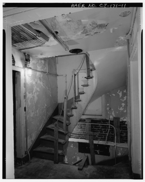 Black and white image of a worn staircase with peeling walls and exposed brick, illuminated by a wall-mounted light.