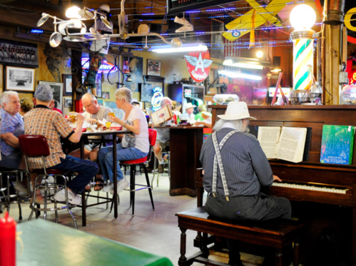 A lively diner scene with patrons eating and chatting, while a man in a hat plays piano in the foreground.