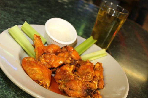 A plate of spicy chicken wings with celery sticks, a small bowl of dipping sauce, and a glass of beverage.
