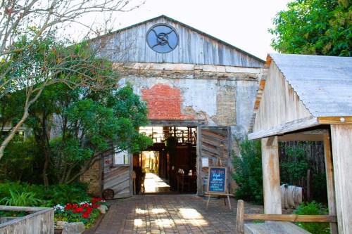A rustic barn entrance with greenery, weathered wood, and a chalkboard sign, inviting visitors inside.