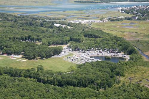 Aerial view of a green landscape with a campground, pond, and nearby waterway, surrounded by trees and open fields.