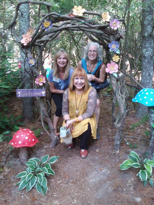 Three women pose together under a floral archway in a forest, surrounded by colorful mushrooms and greenery.
