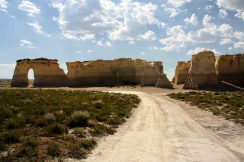 A dirt road leads through a rocky landscape with towering cliffs and a blue sky dotted with clouds.