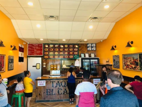 Interior of a vibrant taco restaurant with customers waiting in line, colorful decor, and a menu displayed on the wall.