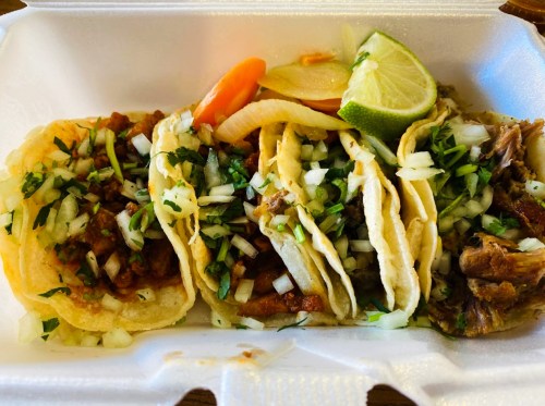 A takeout container with three tacos filled with meat, onions, cilantro, and lime wedges, accompanied by sliced vegetables.