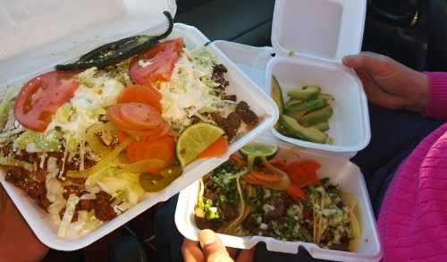 A person holds two takeout containers filled with tacos, meat, vegetables, and garnishes, with a slice of lime.