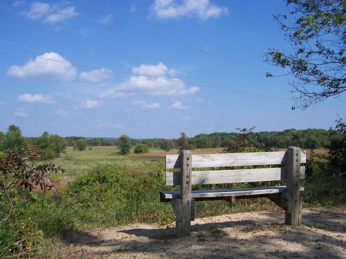 A wooden bench overlooks a grassy field under a blue sky with fluffy clouds and distant trees.