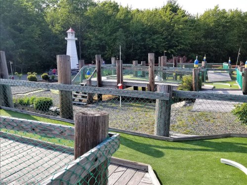A mini-golf course with wooden structures, a lighthouse, and greenery in the background on a sunny day.
