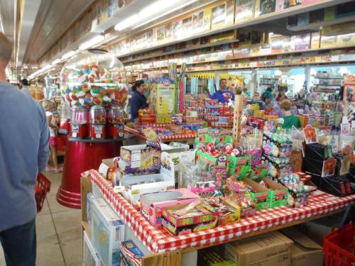 A colorful candy store filled with various sweets, snacks, and a large gumball machine, bustling with customers.