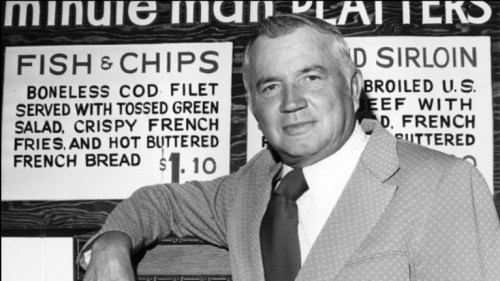 A man in a suit stands beside a menu board featuring fish and chips and sirloin steak options.