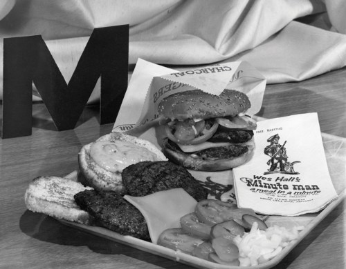A black and white image of a burger platter with a sandwich, sides, and a menu on a wooden table.