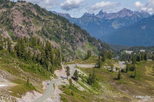 A winding road through lush green mountains, with peaks in the background under a partly cloudy sky.