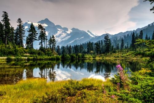 A serene landscape featuring a calm lake, lush greenery, and majestic mountains under a hazy sky.