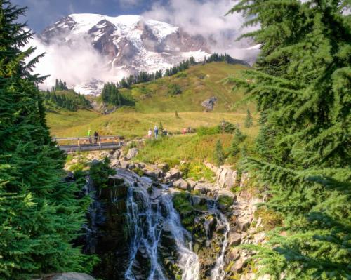 A scenic view of a waterfall surrounded by lush greenery, with snow-capped mountains and clouds in the background.
